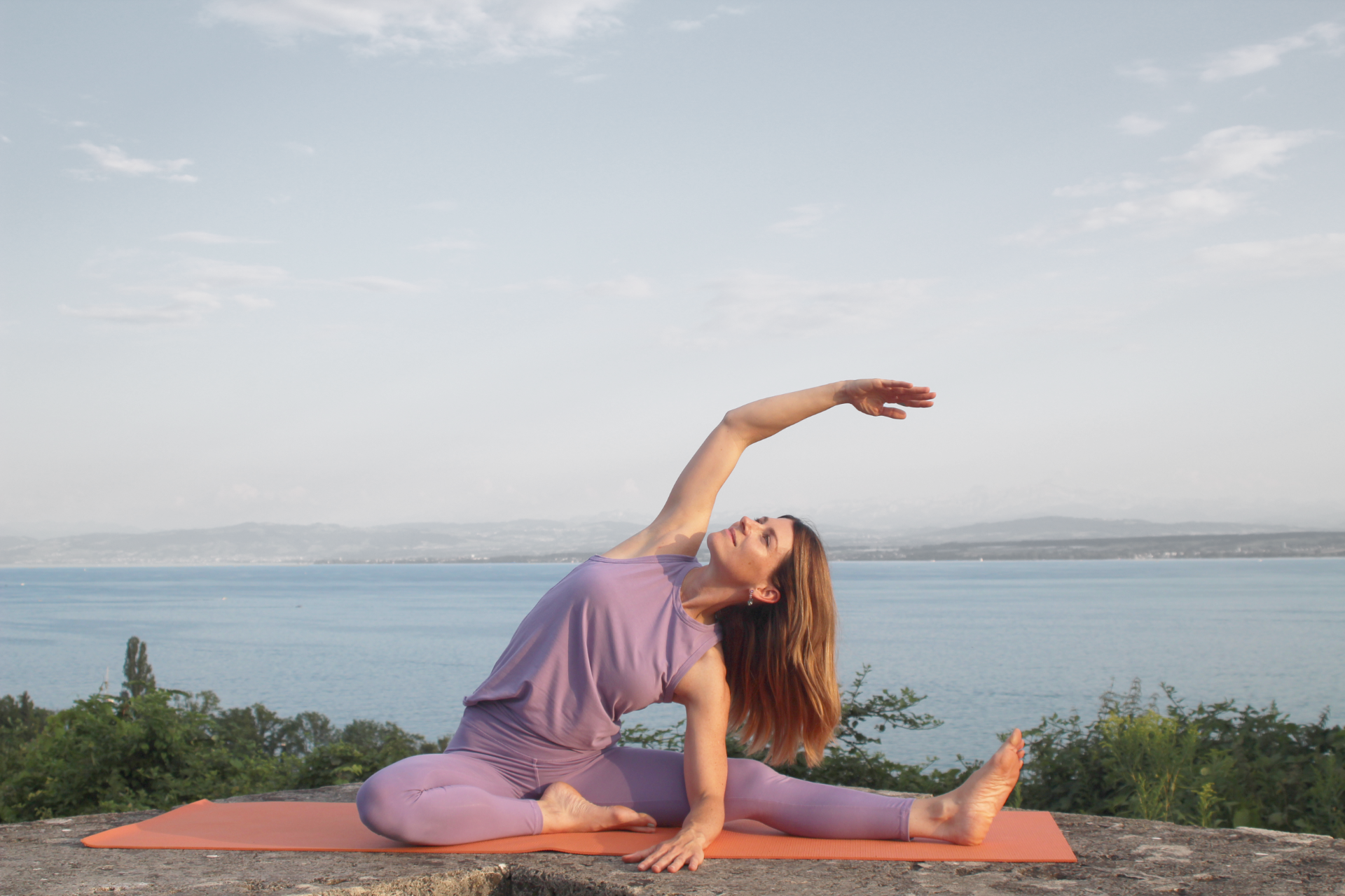 Frau macht auf einer Yogamatte eine Yogaübung, die Asana Janu Sirsasana im Hintergrund ein See und Berge,