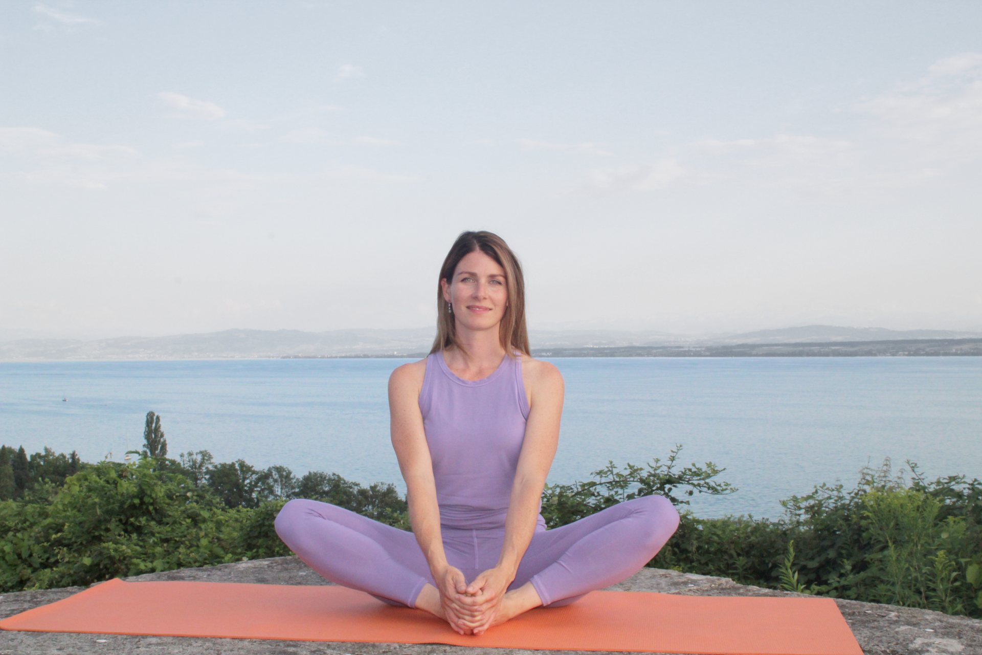 Frau mach auf einer Yogamatte den Schmetterling im Sitzen und schaut und lächelt in die Kamera