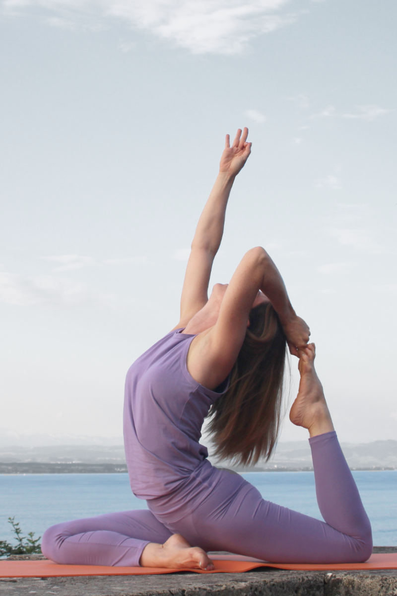 Frau macht auf einer Yogamatte eine Yogaübung, die Asana Taube. Im Hintergrund ist ein See und Himmel zu sehen